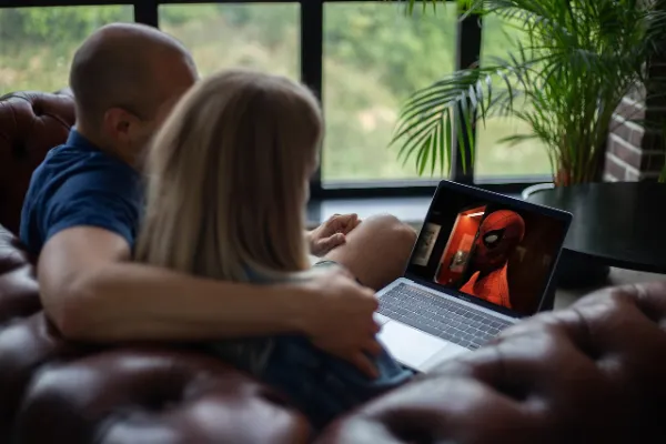 A couple sitting closely on a brown leather couch, watching Spider-Man on a laptop, with a green plant and window in the background.
