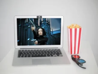 A silver laptop displaying an action movie scene with a woman holding two guns, next to a red and white striped popcorn box and 3D glasses, all on a white surface.