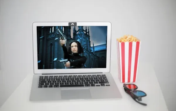 A silver laptop displaying an action movie scene with a woman holding two guns, next to a red and white striped popcorn box and 3D glasses, all on a white surface.