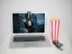 A silver laptop displaying an action movie scene with a woman holding two guns, next to a red and white striped popcorn box and 3D glasses, all on a white surface.