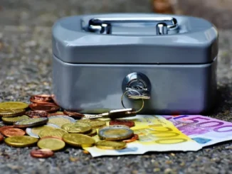 A grey metal cash box with keys in the lock, surrounded by a variety of Euro coins and a 500 Euro banknote on a textured grey surface, illustrating the concept of money.