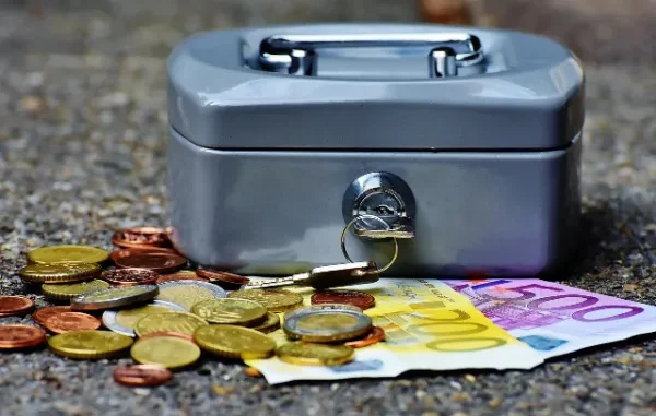 A grey metal cash box with keys in the lock, surrounded by a variety of Euro coins and a 500 Euro banknote on a textured grey surface, illustrating the concept of money.