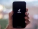 A hand holding a modern smartphone with the TikTok logo and name displayed on a black screen, symbolizing a platform where viral trends and keywords originate.
