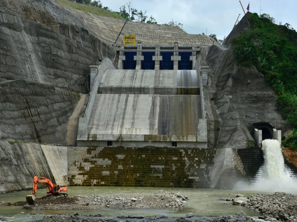 A vast, blue man-made lake or reservoir is held back by a huge, stepped concrete wall of a dam, framed by lush green tropical hills, symbolizing robust infrastructure and water resource management for a resilient community.