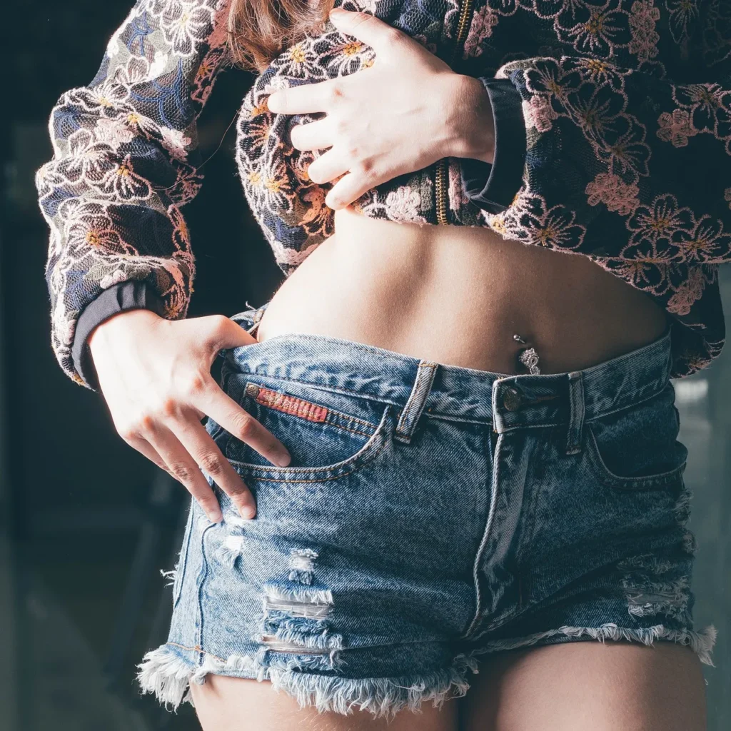 A close-up, cropped image of a young woman lifting her shirt to expose her flat midriff and navel piercing while wearing distressed denim shorts, symbolizing the focus on the body's core and appearance in the discussion of body types.