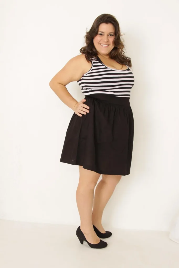 A full-body studio portrait of a smiling woman with curly hair, wearing a striped tank top and black skirt, standing with her hand on her hip, symbolizing diverse body types and the discussion of weight categorization.