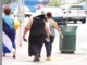 Three women of varying body types and sizes walk away from the camera on a sunny sidewalk in an urban setting, illustrating different body compositions and the topic of weight and stature.