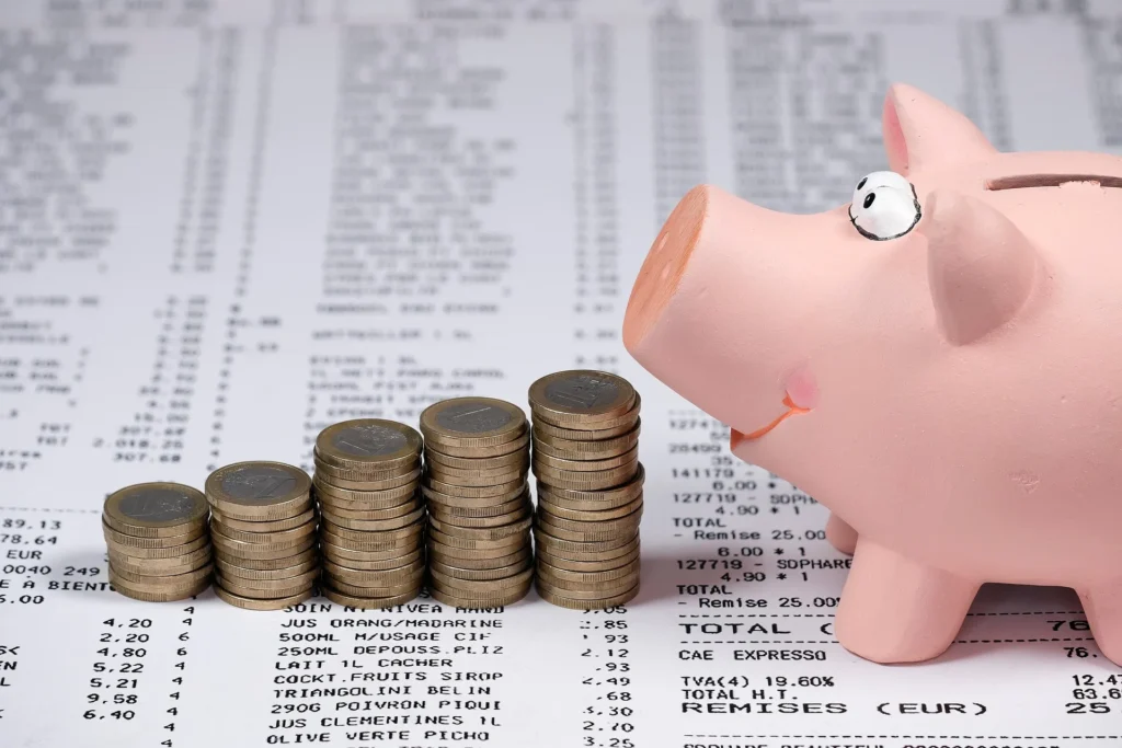 A pink piggy bank tilted upward as if gasping or shouting, positioned next to five stacks of Euro coins arranged in an increasing bar graph style. The scene is set against a background of blurred grocery receipts filled with rising prices, symbolizing the financial stress and psychological impact of inflation on personal savings.