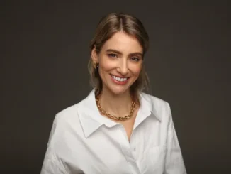 A smiling woman wearing a crisp white button-down shirt and a chunky gold link necklace against a neutral gray background.