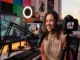 A smiling female content creator with curly hair wearing a headset, sitting in front of a professional streaming setup with a ring light, microphone, gaming monitor, and DSLR camera.