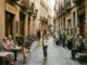 A busy cobblestone street in a European city, featuring outdoor cafe seating, a bookstore, and people walking and relaxing in a relaxed, authentic atmosphere.